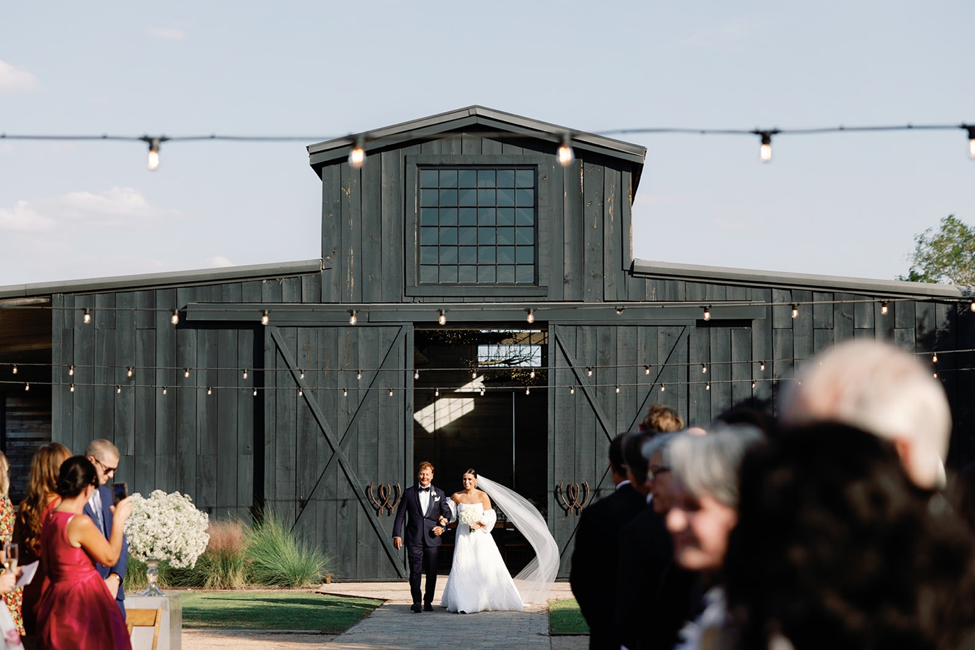 Bride walking down the aisle at Two Wishes Ranch with the barn in the background, walking next to her father