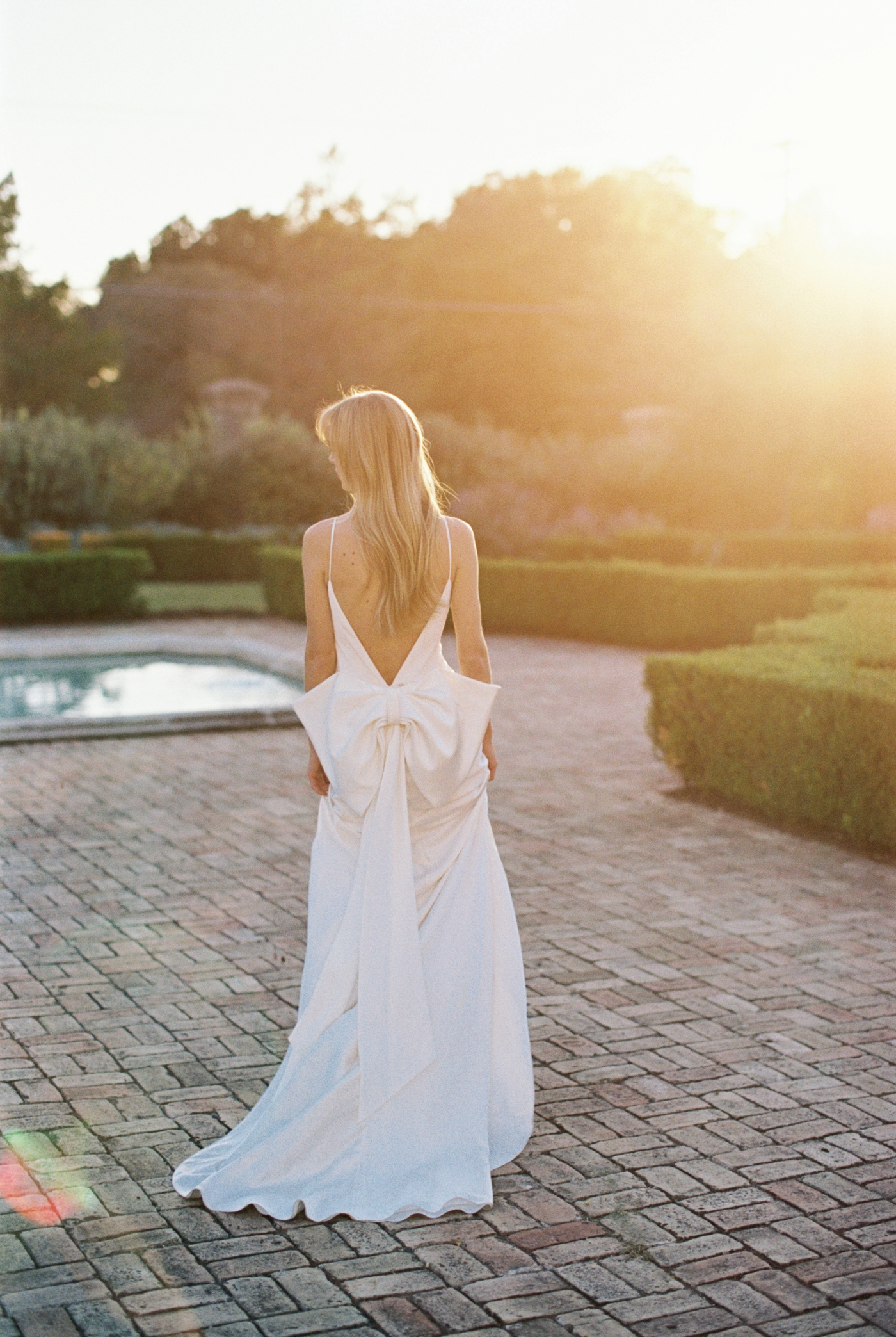 Bride in satin Halfpenny London gown with low back and bow walking with fiancé through garden at Commodore Perry Estate in Austin for their engagement session